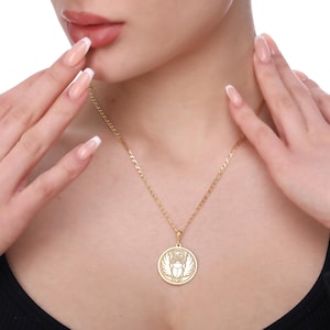 A woman wears a round Skarabeus pendant on a fine link chain just above her chest while lightly touching her face with manicured hands. The engraved scarab beetle with wings and rays draws inspiration from ancient Egyptian amulets and symbolizes rebirth and protection. This medallion necklace comes in solid 14K gold, gold vermeil or 925 sterling silver with gold or rose gold plating. Chain lengths are 16–24 inches and the back can be personalized with initials, dates or affirmations.