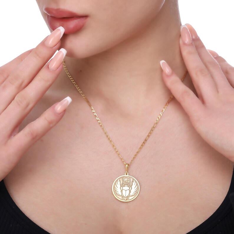 A woman wears a round Skarabeus pendant on a fine link chain just above her chest while lightly touching her face with manicured hands. The engraved scarab beetle with wings and rays draws inspiration from ancient Egyptian amulets and symbolizes rebirth and protection. This medallion necklace comes in solid 14K gold, gold vermeil or 925 sterling silver with gold or rose gold plating. Chain lengths are 16–24 inches and the back can be personalized with initials, dates or affirmations.