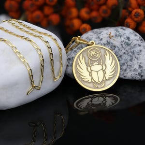 Round medallion necklace with an embossed scarab beetle motif resting on smooth white speckled stones, accompanied by small bright orange berries. The pendant's detailed scarab design is inspired by ancient Egyptian mythology symbolizing rebirth, transformation and protection. Available in solid 14K gold, gold vermeil or 925 sterling silver with gold or rose gold plating. Comes with an adjustable link chain in lengths 16–24 inches and can be personalised on the back with names, dates or affirma
