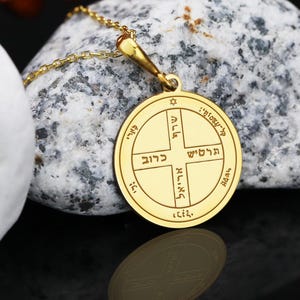 May include: A gold-colored pendant necklace with a circular charm featuring Hebrew text and a star. The pendant hangs on a delicate gold chain. The charm is resting on a gray and white stone.