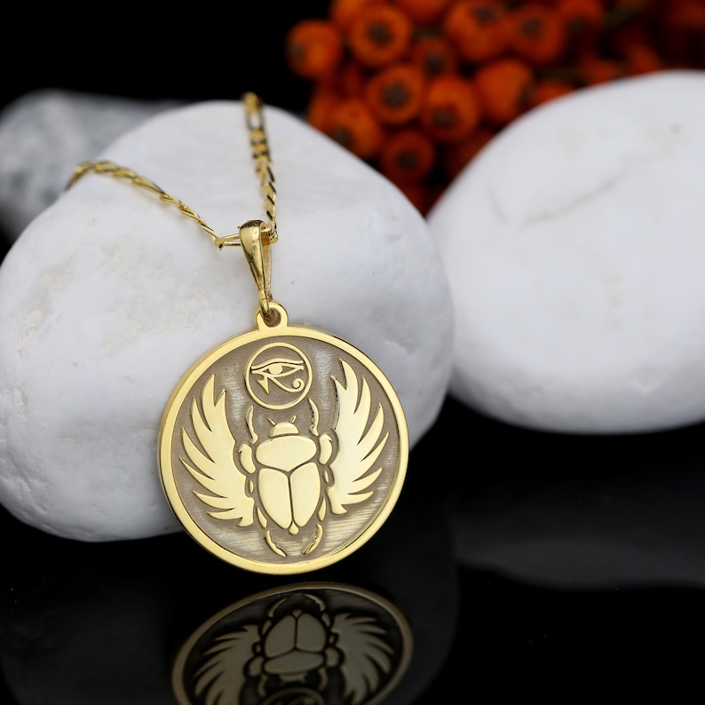 K&ouml;nnte beinhalten: Goldfarbene Anh&auml;ngerkette mit &auml;gyptischem Skarab&auml;us-Design. Der runde Anh&auml;nger zeigt einen detaillierten K&auml;fer mit Fl&uuml;geln und einem Horusauge-Symbol. Die Halskette wird auf einem wei&szlig;en Stein vor schwarzem Hintergrund pr&auml;sentiert.