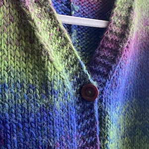 Könnte beinhalten: Nahaufnahme eines gestrickten Cardigans mit V-Ausschnitt und Knopfleiste. Der Cardigan ist aus weichem, flauschigem Garn in Grün-, Blau- und Lilatönen gefertigt.