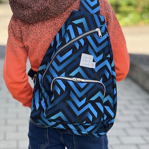 Könnte beinhalten: Ein blau-schwarzer Rucksack mit geometrischem Muster, einem einzelnen Schultergurt, einem großen Hauptfach mit silbernem Reißverschluss und einer kleineren Vordertasche. Ein weißes rechteckiges Etikett mit schwarzem Text ist vorne aufgenäht.