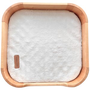 May include: A light-colored wooden pet bed with a plush, white, rectangular cushion. The bed has rounded corners and a small, rectangular tag that reads "ERGO PURRCH".