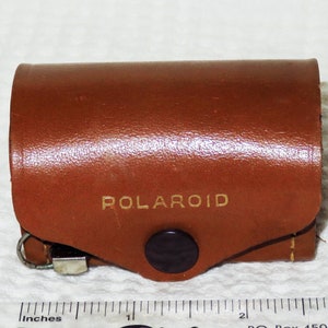 Puede incluir: Una funda de cuero marrón con un logotipo "POLAROID" en relieve dorado y un botón negro.
