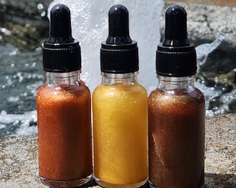May include: Three glass dropper bottles filled with shimmering liquid in shades of bronze, gold, and brown. Each bottle has a black cap and dropper. The bottles are arranged in a row against a blurred background of a fountain.