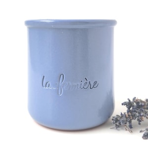 May include: A light blue ceramic jar with the embossed text "la fermière". The jar has a cylindrical shape and a slightly flared rim. Dried lavender sprigs are next to the jar. The background is white.