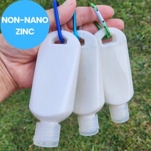 May include: Three white, rectangular bottles with clear caps are attached to colorful carabiners. The bottles are filled with a white substance and the text "NON-NANO ZINC" is in a blue circle in the upper left corner.