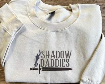 May include: A light gray sweatshirt with the text "SHADOW DADDIES" in a stylized font, accompanied by a flame and sword graphic. The sweatshirt is folded on a wooden surface.