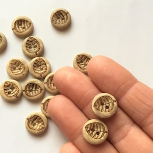 May include: A collection of wooden buttons with a leaf design. The buttons are light brown with a darker brown leaf design.