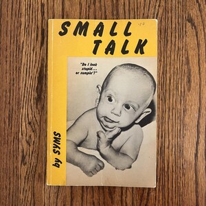 May include: A yellow and white book cover with the title "SMALL TALK" in black bold letters. The cover features a black and white photo of a baby with a thoughtful expression, and the text "Do I look stupid... or sumpin'?" is printed below the photo. The author's name, "by SYMS", is printed vertically on the left side of the cover.