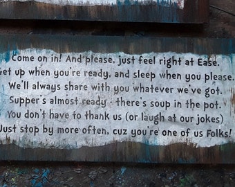 May include: A weathered wooden sign with white painted text that reads: "Come on in! And please, just feel right at Ease. Get up when you're ready, and sleep when you please. We'll always share with you whatever we've got. Supper's almost ready - there's soup in the pot. You don't have to thank us (or laugh at our jokes) Just stop by more often, cuz you're one of us Folks!"