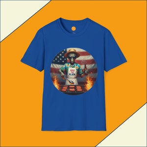 Puede incluir: Camiseta azul real con un gráfico circular de un extraterrestre asando perritos calientes frente a una bandera estadounidense. El extraterrestre lleva una camisa hawaiana, un delantal con el texto "Kiss the Alien" y un sombrero de paja.