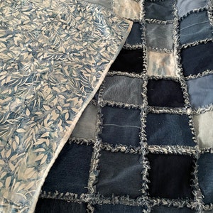 May include: A patchwork quilt made from denim fabric with frayed edges. The quilt is a mix of light and dark blue denim squares. A white fabric with a blue leaf pattern is visible in the background.