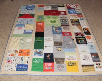 May include: A patchwork quilt made from baby onesies and t-shirts. The quilt is beige with a blue border. The onesies and t-shirts have various designs and text, including "Heartbreaker", "Rehoboth", "Yes", "Wild Child", "King of the Beach", "Mom", "Tough Like Dad", "Grandma Rocks", and "Cool Like Auntie".