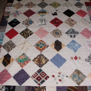 May include: A white quilt with a diamond pattern made from various fabrics, including patterned ties, floral prints, and animal prints. The quilt has a red border.