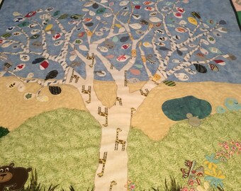 May include: A white fabric tree with a brown bear, giraffes, and green grass at the base. The tree is decorated with colorful fabric leaves and butterflies. The background is a light blue sky.