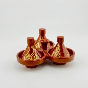 May include: A set of three small, terracotta-colored ceramic tagines, each with a conical lid and a circular base. The tagines are decorated with yellow floral patterns. The set is designed to hold multiple dishes.