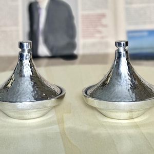 Peut inclure: Deux tajines en métal argenté, martelé, avec couvercles coniques et bases rondes. Les pots sont présentés sur une surface en bois clair. L'arrière-plan est flou, mais semble être un journal et une photo encadrée.