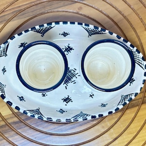 May include: Oval ceramic serving dish with two cup holders. The dish is white with a blue rim and decorative blue geometric patterns. The dish is sitting on a wooden surface.
