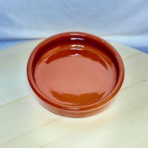 May include: A small, round, terracotta-coloured ceramic bowl with a glossy finish. The bowl has a shallow depth and a slightly raised rim. It sits on a light-coloured wooden surface.