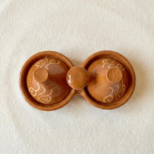 May include: A brown ceramic salt and pepper shaker set. The set features two round containers connected by a bridge, each with a conical lid and a decorative swirl pattern. The set is made of earthenware.