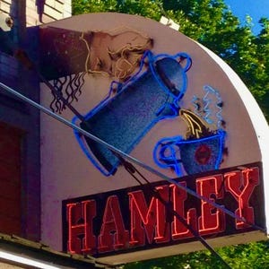 May include: A neon sign with the word "HAMLEY" in red letters. The sign is lit up and has a blue and purple image of a coffee pot pouring coffee into a cup.