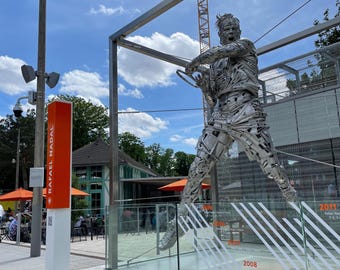 Könnte beinhalten: Eine große, silberne Skulptur eines Tennisspielers im Schwung, umgeben von einem Glasgehäuse. Die Skulptur besteht aus Metallschrott und ist auf einem Betonsockel montiert. Die Skulptur befindet sich vor einem Gebäude mit einem roten Schild, auf dem "Rafael Nadal" steht.