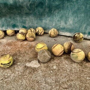 May include: A group of yellow tennis balls with some showing signs of wear and tear, scattered on a rough, brown surface. Some of the balls have the word "Babolat" printed on them.