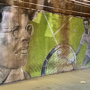 May include: A mural on a concrete wall depicts two people playing tennis. The person on the left is a woman with long brown hair wearing glasses. The person on the right is a man with short brown hair wearing a white shirt and shorts. The mural is painted in shades of green, brown, and white.