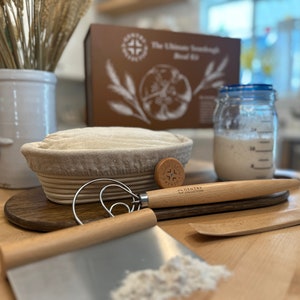 May include: A close-up of a sourdough bread baking kit. The kit includes a proofing basket, a dough scraper, a wooden rolling pin, a metal dough whisk, and a jar of sourdough starter. The kit is on a wooden table with a bag of flour in the background.