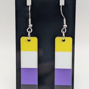 May include: Non-binary pride flag earrings. These earrings feature a rectangular design with a yellow, white, purple, and black color scheme. The earrings are suspended from silver-colored hooks.