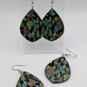 May include: Teardrop-shaped earrings with a Bigfoot and forest design. The earrings feature a black background with green, blue, and brown trees and a Bigfoot figure. The earrings are displayed on a clear stand.