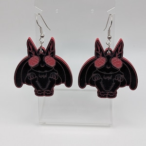 May include: A pair of dangle earrings featuring a black bat-like creature with large, textured red eyes and red outlines. The earrings are suspended from silver-toned metal hooks and display a detailed, symmetrical design of the creature.