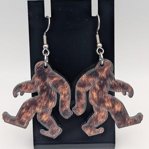 May include: A pair of Bigfoot-shaped earrings. The earrings feature a brown and tan fur pattern on a black outline. The earrings hang from silver-colored fish hook ear wires. The earrings are displayed on a black stand.