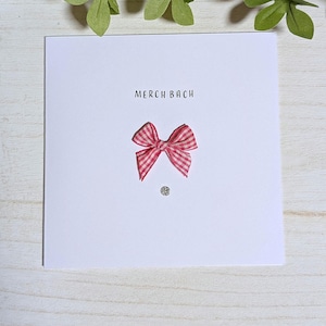 May include: A white greeting card with the text "MERCH BACH" in black. A pink and white gingham bow is centered below the text, with a small silver embellishment beneath it. The card is on a light wood surface.