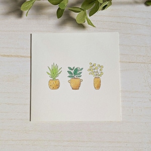 Op de afbeelding: Waterverfillustratie van drie planten in potten. De planten staan in geel-oranje potten met verschillende bladvormen en -texturen. Het kunstwerk staat op een wit vierkant papier, met een houten achtergrond en een takje groene bladeren bovenaan.