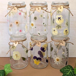 Op de afbeelding: Zes heldere glazen potten versierd met gedroogde bloemen en touw. Elke pot is versierd met een touwstrik en een klein houten hart. De potten zijn gevuld met diverse gedroogde bloemen, waaronder madeliefjes en viooltjes.