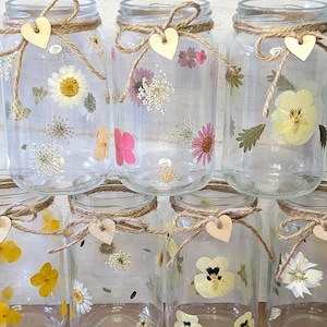 May include: Six clear glass mason jars decorated with various pressed flowers in shades of yellow, pink, and white. Each jar features a natural jute twine bow tied around the neck, accented with a small, light-colored wooden heart charm.