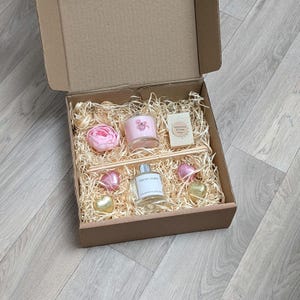 Könnte beinhalten: Eine braune Geschenkbox mit verschiedenen Artikeln. Darin befinden sich eine rosa Kerze, eine Flasche "Fresh Linen" Duft, ein Stück Seife, eine rosa Rose und herzförmige Pralinen, alles auf Stroh gebettet.
