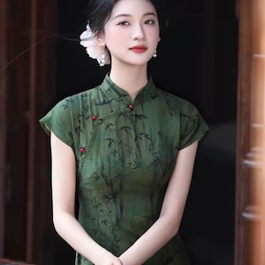 Könnte beinhalten: Ein dunkelgrünes Cheongsam-Kleid mit schwarzem Bambusblattmuster, Stehkragen und kleinen roten, kugelförmigen Knöpfen entlang des seitlichen Verschlusses. Das Kleidungsstück hat kurze Ärmel und eine körperbetonte Passform im traditionellen Stil.