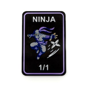 May include: A rectangular black patch with a light purple border featuring the white text NINJA at the top and 1/1 at the bottom. In the centre, a stylised blue and white illustration depicts a ninja in a dynamic leaping pose throwing a shuriken.