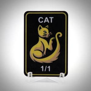 Peut inclure: Un autocollant rectangulaire noir aux coins arrondis, avec une illustration de chat jaune et le mot "CAT" en haut. Le numéro "1/1" est imprimé en bas. L'autocollant est présenté sur un support en plastique transparent.