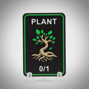 May include: A black rectangular card with a green border and the word "PLANT" in white. A stylized tree illustration with green leaves and a beige trunk and roots is centered below the word. The card reads "0/1" at the bottom.