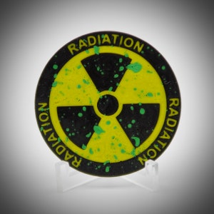 May include: A black and yellow radiation symbol with the word "RADIATION" in yellow text around the edge. The symbol has green splatter details and is displayed on a clear stand.