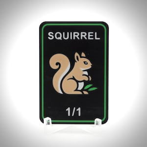 May include: A black rectangular card with a green border displays the word "SQUIRREL" in white. A tan squirrel illustration is below the text, with "1/1" at the bottom. The card is presented on a clear stand.