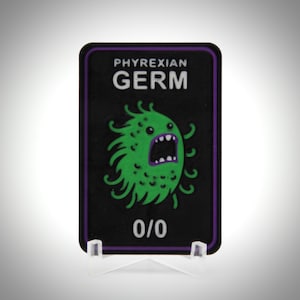 May include: A black rectangular card with the words "PHYREXIAN GERM" in white text and a green cartoon germ with an open mouth. The card has a purple border and the numbers "0/0" at the bottom.