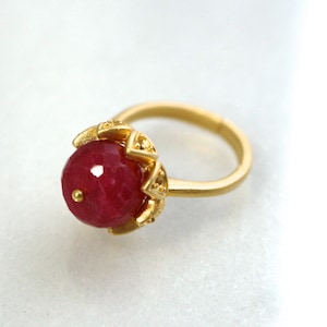 May include: A gold ring with a large, round, red gemstone set in a gold flower-shaped setting.