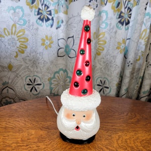 May include: A white ceramic Santa Claus head with a red and white hat. The hat has green and red jewels around it. The Santa Claus head has a white beard and a red mouth.