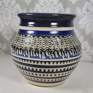 May include: A handcrafted ceramic vase with a dark blue rim and a cream-colored base. The vase features a series of blue and cream-colored horizontal stripes, with a wavy pattern in the middle. The vase is approximately 7 inches tall and 6 inches wide.